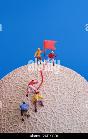 Giocattoli in miniatura - un gruppo di alpinisti che raggiungono la cima. Rock melone frutta come base. Foto Stock