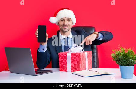 uomo felice indossa il cappello rosso di babbo natale e gli occhiali da festa contengono la confezione regalo, presentando lo smartphone Foto Stock