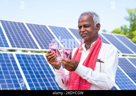 Buon contadino che conta denaro o banconote di fronte al pannello solare presso il terreno agricolo - concetto di approvazione del prestito, sovvenzione bancaria e risparmio o investimento. Foto Stock