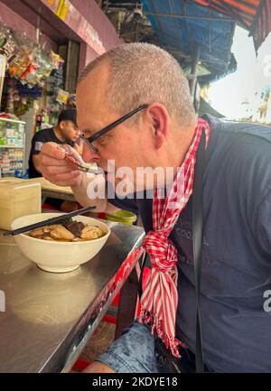 Un turista straniero di mezza età mangia una ciotola di Bun rieu, una forma di zuppa vietnamita di noodle, nel centro di Phnom Penh, Cambogia. Foto Stock