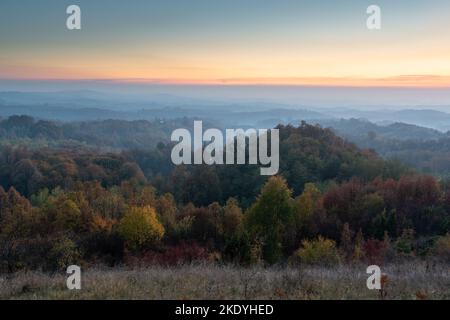 Paesaggio rurale durante il crepuscolo in autunno, campagna collinare con lussureggianti foreste colorate e arancione bagliore nel cielo Foto Stock
