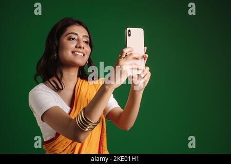 Giovane donna indiana con sari che prende selfie sul telefono cellulare isolato su sfondo verde Foto Stock