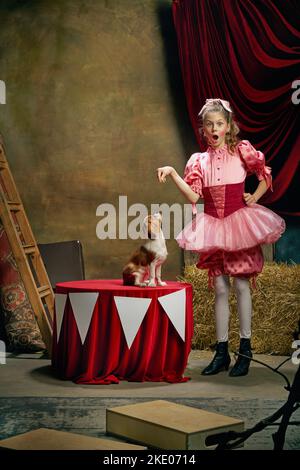 Allenatore di circo. Ritratto di piccola bella ragazza carina in vestito festivo addestramento divertente doggy al circo vintage. Vacanze, sogni, arte, moda Foto Stock