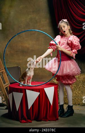 Allenatore di circo. Ritratto di piccola bella ragazza carina in vestito festivo addestramento divertente doggy al circo vintage. Vacanze, sogni, arte, moda Foto Stock