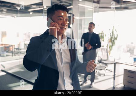 Occupato uomo d'affari asiatico parlare telefono con il cliente mentre in piedi in ufficio sullo sfondo di un collega Foto Stock