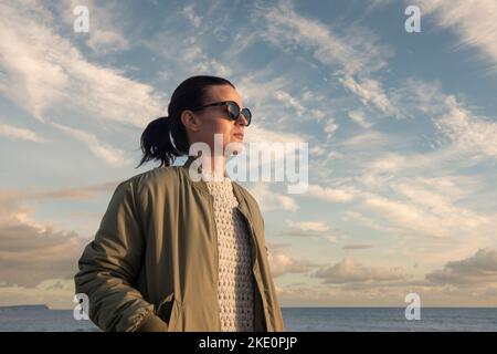 Ritratto di una donna che indossa occhiali da sole all'aperto vicino al mare con un cielo spettacolare Foto Stock