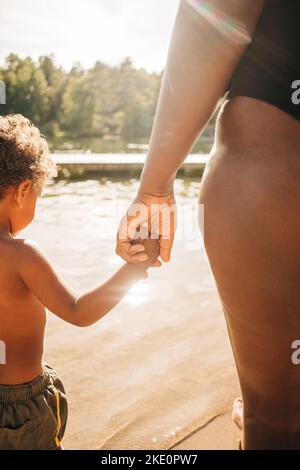 Donna che tiene la mano del figlio mentre si trova vicino al lago nella giornata di sole Foto Stock