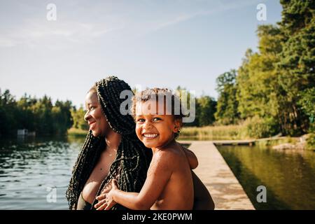 Donna sorridente che trasporta il figlio eccitato al lago durante la vacanza Foto Stock