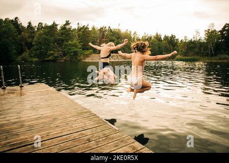Amici donne che indossano bikini che saltano in lago con le braccia allungate durante le vacanze Foto Stock