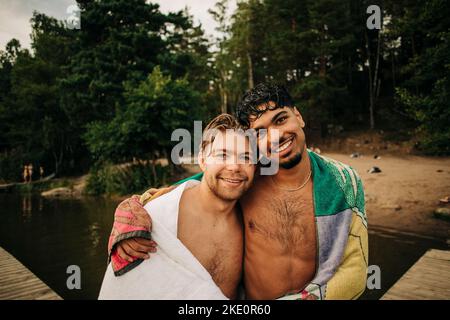 Ritratto di giovane uomo con braccio intorno a un amico maschio che indossa asciugamano durante le vacanze Foto Stock