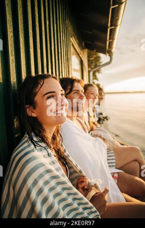 Felice donna contemplativa che indossa asciugamano seduto con gli amici vicino al cottage Foto Stock