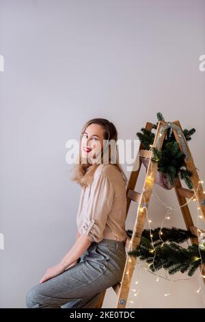 Ritratto di donna moderna di mezza età a scala di legno, decorato con rami di abete, ghirland per Natale. Posiziona per testo, parete luminosa. Scandinavian st Foto Stock