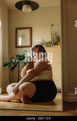 Giovane donna che prende il selfie attraverso lo smartphone mentre si siede a gambe incrociate contro lo specchio a casa Foto Stock