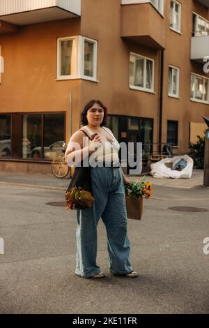 Ritratto di giovane donna fiduciosa con la borsa della spesa in piedi contro la costruzione Foto Stock