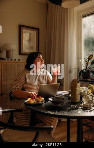 Giovane donna che tiene il bicchiere mentre si siede con il computer portatile al tavolo Foto Stock