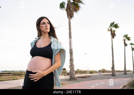 giovane donna incinta con lunghi capelli castani indossa abbigliamento sportivo e fa attività fisica delicata come stretching e camminare su una pista e ha anche tempo Foto Stock