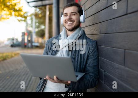 giovane uomo elegante che ascolta la musica con le cuffie e che gli copre gli occhi con un computer portatile nelle mani all'esterno Foto Stock