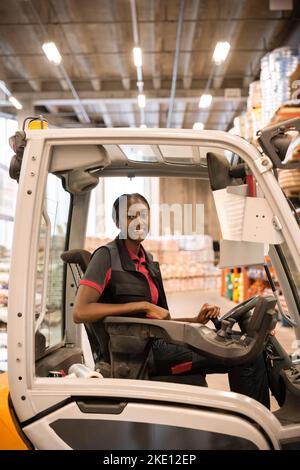 Vista laterale di una lavoratrice di magazzino seduta in un carrello elevatore a forche in magazzino Foto Stock
