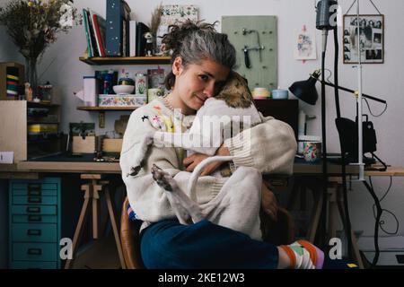 Ritratto di donna che indossa un maglione che abbraccia il cane mentre si siede sulla sedia a casa Foto Stock