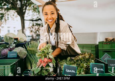 Ritratto di felice proprietario femmina che tiene verdure vicino alle casse in stalla al mercato Foto Stock