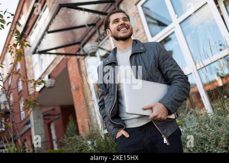 sottoscrittore maschio all'ingresso dell'edificio degli uffici con un sorriso con un computer portatile in mano Foto Stock