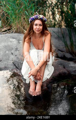 La sposa siede sulla riva del fiume e guarda la fotocamera Foto Stock