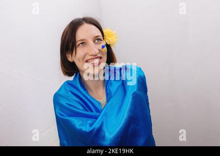 La bandiera dell'Ucraina è dipinta sulla guancia della ragazza con vernice blu e gialla. Fiore nei suoi capelli. La donna sorride sinceramente e guarda via. Giornata dell'indipendenza dell'Ucraina. Spazio di copia. Foto Stock