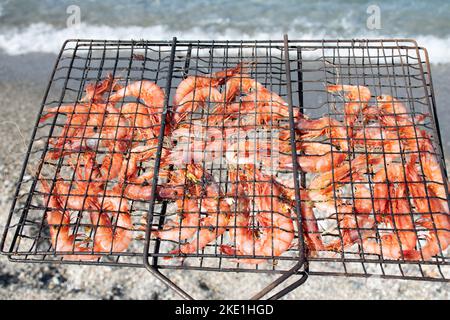 Gamberetti grigliati in una griglia barbecue tenuta di fronte al mare con ciottoli di spiaggia sfocati e un'onda sullo sfondo Foto Stock