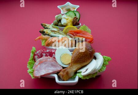 Piatto in forma di albero di Natale con cibo proteico - carne, pesce, formaggi, noci, ecc sfondo rosso. Il concetto di dieta del keto tratta per il holi Foto Stock