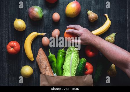 Una mano che raccoglie una frutta da un sacchetto di shopping riempito di verdure, frutta, uova e pane su un fondo di legno. Scelte alimentari sane. Superma online Foto Stock