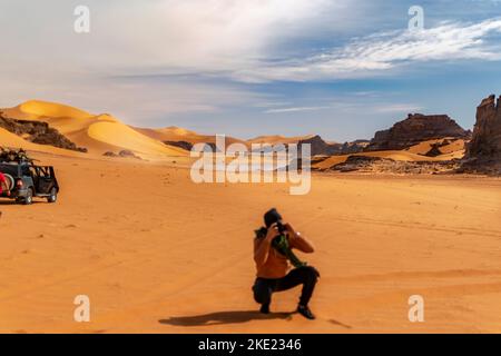 Fotografo sfocato accovacciato e in possesso di una fotocamera DSLR. Tadrart Rouge Sahara Desert dune di sabbia e montagna rocciosa. 4WD veicolo parcheggiato sul reg. Foto Stock