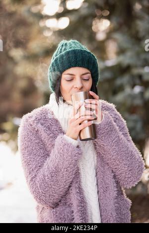 Primo piano ritratto della donna che cammina il giorno d'inverno, tenendo in mano tazza di viaggio in acciaio inox con caffè caldo. Bottiglia d'acqua riutilizzabile. Rifiutare, ridurre, riciclare a. Foto Stock