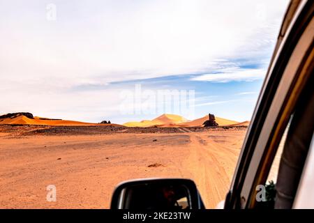 Vista dei passeggeri posteriori dal finestrino esterno di un'auto in movimento nel deserto del Sahara. Fuoristrada reg, dune di sabbia, mesa rocciosa e un cielo blu coperto a Tassili n'Ajjer. Foto Stock
