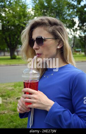 Ragazza bionda attraente in occhiali da sole beve un cocktail effervescente attraverso una paglia Foto Stock