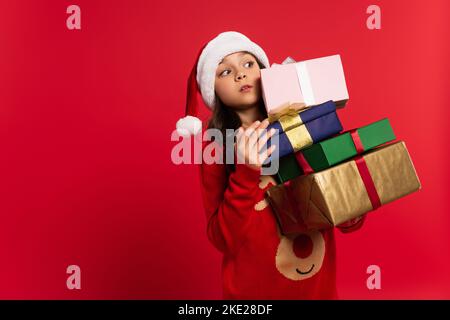 Ragazza sorpresa in cappello di santa e maglione che tiene mazzo di regali di Natale isolato sul rosso Foto Stock