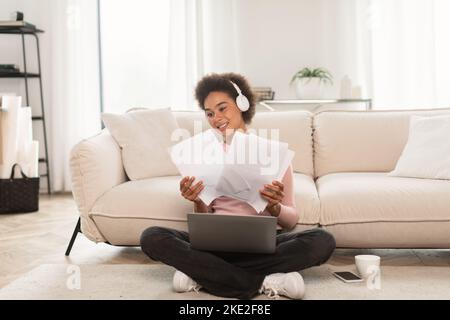 Felice millenario afroamericana femminile in cuffie wireless ha videochiamata sul computer portatile, funziona con i documenti Foto Stock