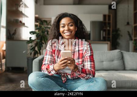Allegra giovane donna nera in stile casual digitando sullo smartphone, chattando e utilizzando la nuova app Foto Stock