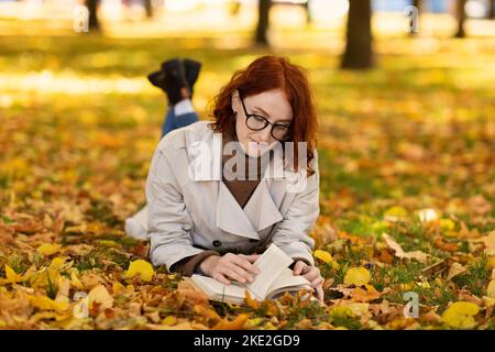 Donna millenaria caucasica concentrata seria con capelli rossi in impermeabile e occhiali giace sul terreno Foto Stock