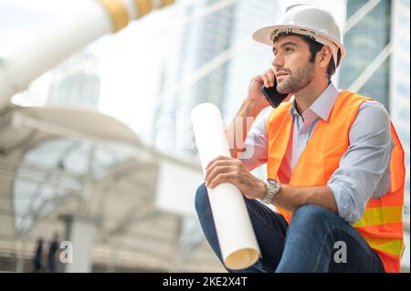 Gli ingegneri del Caucaso usano uno smartphone per parlare, indossando un giubbotto arancione e un grande elmetto, e l'altra mano che tiene la pianta bianca del pavimento. Foto Stock