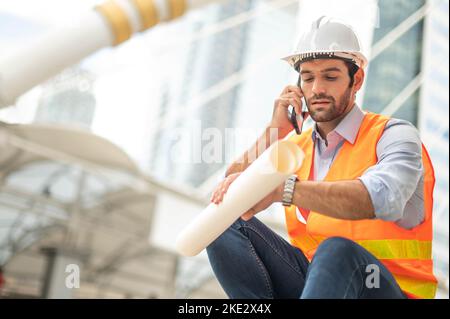 Gli ingegneri del Caucaso usano uno smartphone per parlare, indossando un giubbotto arancione e un grande elmetto, e l'altra mano che tiene la pianta bianca del pavimento. Foto Stock