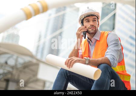 Gli ingegneri del Caucaso usano uno smartphone per parlare, indossando un giubbotto arancione e un grande elmetto, e l'altra mano che tiene la pianta bianca del pavimento. Foto Stock