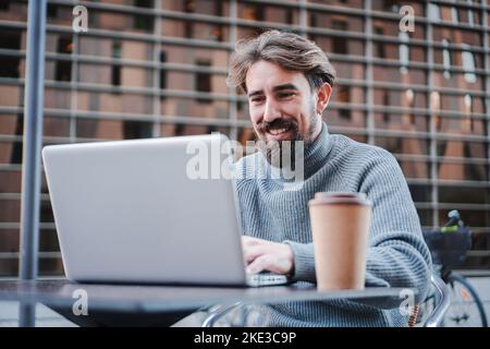 Giovane uomo d'affari sorridente che usa il laptop per lavorare in remoto dalla terrazza del caffè, Ritratto di un freelance di successo o di uno studente che lavora su un nuovo progetto Foto Stock