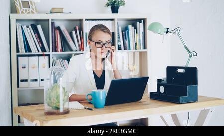 Una giovane donna sicura che lavora in azienda parla al telefono cellulare, usa il notebook e prende appunti sul notebook seduto alla scrivania sul posto di lavoro. Business, ufficio moderno e concetto di persone. Foto Stock