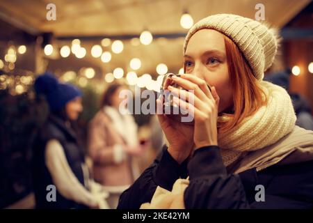 Focus su giovane ragazza sorridente bella in abiti invernali bere bevande calde e sognare. Trascorrere del tempo con gli amici alla fiera invernale a sera. Foto Stock