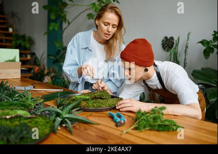 Fiorista donna che insegna giovane uomo hipster decoratore a studio arte artigianale Foto Stock