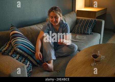 Giovane donna felice usando la lima del chiodo mentre si dà pedicure nel paese. Donna che applica smalto unghie sulle sue unghie a casa. Donna seduta sul divano pa Foto Stock