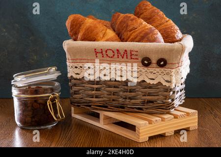 Quattro croissant in un cestino di vimini. C'è un vasetto di uva passa sul tavolo. Composizione su sfondo blu. Foto Stock