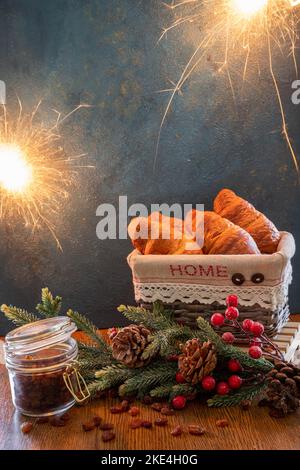 Quattro croissant in un cestino di vimini. Sulla tavola si trova un ramo di abete rosso con coni e bacche, accanto ad esso si trova un vasetto di uva passa. Bengala si accende su una ba blu Foto Stock