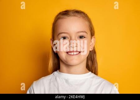 Ritratto di piccola bella ragazza, bambino in posa T-shirt bianca, sorridente alla macchina fotografica isolato su sfondo giallo studio Foto Stock