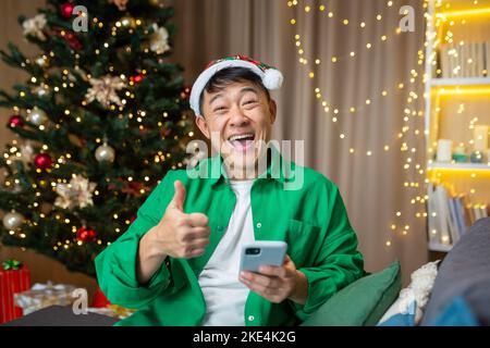 Un giovane bell'uomo asiatico tiene un telefono nelle sue mani, è felice, ha ricevuto buone notizie alla vigilia di Natale e di Capodanno. Super punti, seduti sul divano vicino all'albero di Natale. Foto Stock